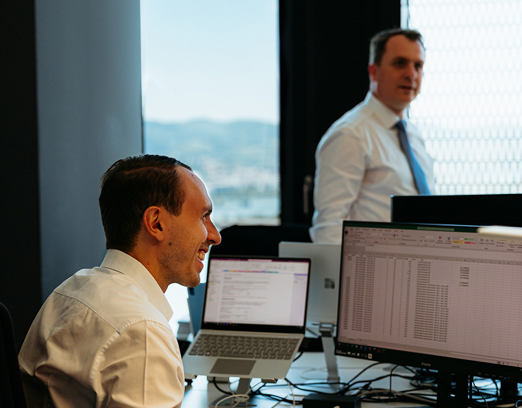 Two men in business attire working at desks with laptops and monitors displaying spreadsheets in a modern office. One man is seated and smiling; the other is standing in the background.