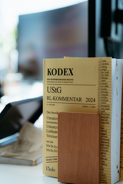 A yellow-covered German legal commentary book titled KODEX UStG RL-KOMMENTAR 2024 stands upright on a desk beside a brown notepad.