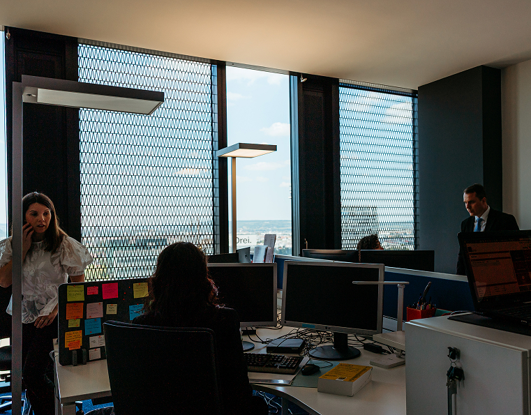 Four people work in a modern office with large windows covered by metal mesh; computer monitors, desks, and sticky notes are visible.